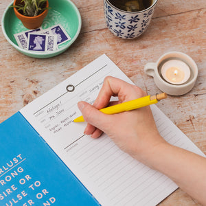 A hand holding a yellow pen writes in a travel logbook on a rustic wooden table, surrounded by cozy props including a blue-and-white floral mug, a small potted succulent, postage stamps, and a lit candle.