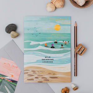 A beach-themed illustrated logbook titled 'WILD SWIMMING LOGBOOK' is displayed in a styled flat-lay arrangement with shells, a pencil, a pencil sharpener, and a card on a light gray surface.