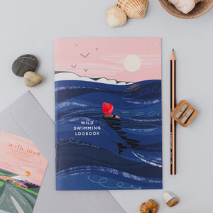 A flat-lay photograph of a 'Wild Swimming Logbook' featuring an illustration of a person swimming on a jet ski with a red heart. The book is styled on a light gray surface with seashells, a pencil, a sharpener, and another card.