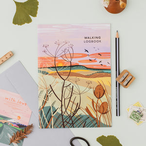 A flat-lay of a 'Walking Logbook' featuring a colorful landscape illustration with wildflowers and birds at sunset. The book is styled on a light gray surface with a matching envelope, a card that says 'with love', a pencil, a wooden pencil sharpener, and other botanical-themed stationery.