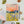 A square illustrated logbook titled 'WALKING LOGBOOK' lies on a light surface, surrounded by stationery and small decorative items. The cover art shows a stylized wildflower landscape with a peach and orange sky, yellow field, blue-purple flowers, and dark green stems.