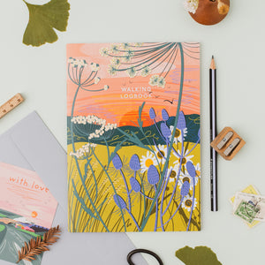 A square illustrated logbook titled 'WALKING LOGBOOK' lies on a light surface, surrounded by stationery and small decorative items. The cover art shows a stylized wildflower landscape with a peach and orange sky, yellow field, blue-purple flowers, and dark green stems.
