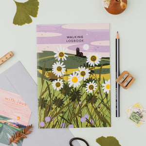 A flat-lay of a 'Walking Logbook' featuring a whimsical landscape illustration with daisies, a cliff edge with a lighthouse, and a purple sky. The book is styled on a light gray surface with a pencil, wooden pencil sharpener, a pink card, and other small decorative items.