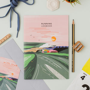 An illustrated running logbook titled 'RUNNING LOGBOOK' lies on a light surface with a matching card reading 'with love' beside it. The cover art shows a pink sky, orange sun, birds, and green rolling hills.