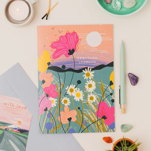 A colorful illustrated gratitude logbook with wildflower artwork and the words 'GRATITUDE LOGBOOK' on the cover, styled on a light surface with a pen, crystals, a candle, and a small plant.