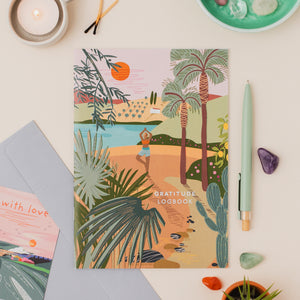 A gratitude logbook with a colorful tropical beach illustration, styled in a flat lay with a mint green pen, crystals, a candle, and a gray envelope on a light beige background.