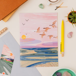 An illustrated reading logbook with a pastel sunset and wild geese design, styled in a flat lay with a yellow pen, small plant, crystals, and other stationery on a light surface.