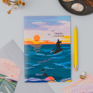 A travel logbook with a colorful illustration of a person paddleboarding on the ocean at sunset, styled in a flat lay with a yellow pen, an envelope, and small cards on a light gray surface.