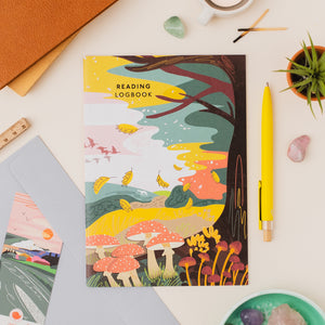A colorful illustrated reading logbook titled 'READING LOGBOOK' is shown in a styled flat-lay with a yellow pen, small plant, crystals, and other stationery on a light neutral background.