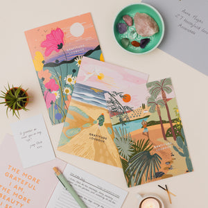 Three illustrated 'Gratitude Logbook' covers are arranged on a light beige surface with a small potted succulent, a mint bowl of stones, a candle, and handwritten notes around them.