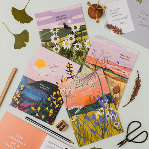 Four illustrated walking logbooks with floral and landscape artwork are arranged on a light surface with stationery props including a pencil, scissors, ruler and a note card.