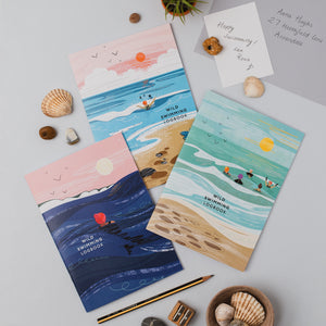 Three illustrated 'Wild Swimming Logbooks' with different cean scenes are arranged on a light gray surface with shells, a pencil, wooden blocks, and a handwritten note.