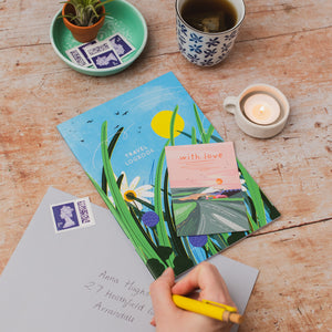 A hand writing on a letter with a yellow pen over a travel logbook and envelope on a rustic wooden table, surrounded by a cup of tea, a lit candle, and a small potted plant. 