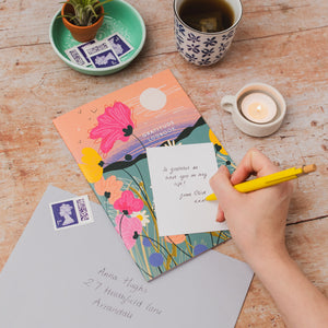 A hand writes on a small white note with a yellow pencil, placed on top of a colorful illustrated postcard. The scene is styled on a wooden table with a cup of tea, a lit candle, a mint dish with small plants, and a gray letter addressed by hand.