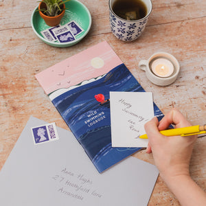 A styled flat-lay of a book titled 'wild swimming logbook' on a rustic wooden table, surrounded by a cup of tea, a candle, a small plant, and a hand writing a letter with a yellow pen.