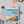 A flat-lay photograph of a 'Wild Swimming Logbook' featuring an illustrated beach and ocean scene. The book is styled on a light gray surface with seashells, a pencil, a wooden pencil sharpener, and another matching card partially visible.
