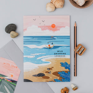 A flat-lay photograph of a 'Wild Swimming Logbook' featuring an illustrated beach and ocean scene. The book is styled on a light gray surface with seashells, a pencil, a wooden pencil sharpener, and another matching card partially visible.