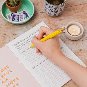 A hand holding a yellow pen writes in a lined journal on a wooden table, surrounded by a blue floral mug, a small potted plant, vintage-style postcards, and a candle.
