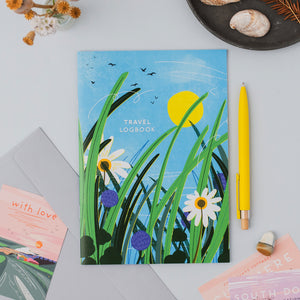 A travel logbook with a colorful illustration of grass and wildflowers and a yellow sun on a blue sky background, styled in a flat lay with a yellow pen, envelope, and postcards on a light gray surface.