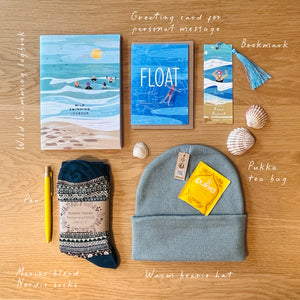 A curated flat lay of multiple small gift items on a wooden surface, including a swimming-themed card, a wild swimming bookmark with tassel, a beanie hat with a pukka tea bag, patterned socks, a pen, and shells. Handwritten labels identify each item.