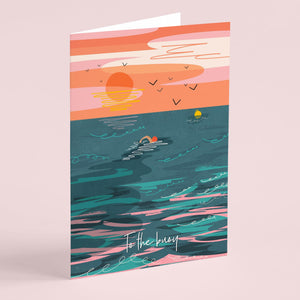 Greeting card with a colourful design of a sunset, ocean, and a wiild swimmer swimming to the buoy