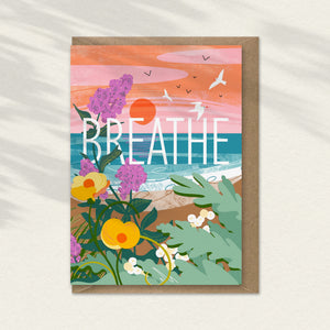 Colourful illustration with 'BREATHE' text with the sea in the background, seagulls flying over and seakale and wild flowers in the foreground. A mindful greeting card illustrated by onneke