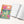 A colorful gratitude logbook with a floral cover design is shown next to an open page of lined writing space on a light beige background.