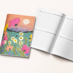 A colorful gratitude logbook with a floral cover design is shown next to an open page of lined writing space on a light beige background.