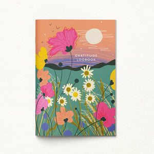 A vertical gratitude logbook with a colorful illustrated cover showing wildflowers, hills, birds, and a sun, photographed against a light textured background.