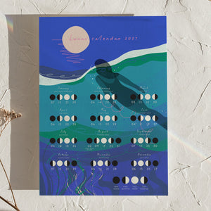 A square illustrated calendar titled 'Luna calendar 2027' leaning against a textured off-white wall. The design features a dark blue night sky with a large pale moon, teal and green wave-like forms, and rows of monthly date blocks with moon phase graphics.
