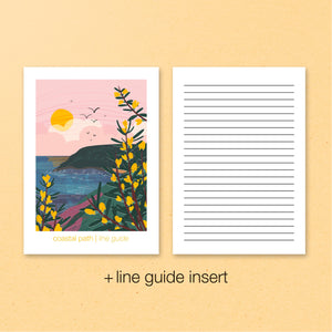 Notebook with a coastal path illustration on one page and a line guide insert on the other, on a beige background.