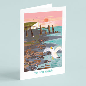 Greeting card with a seagull in the shoreline, a pebble beach and cliffs in the background. Nature illustration on coastal card by onneke