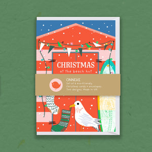 A rectangular printed card set titled "Christmas at the beach hut" with a tan paper band across the center reading "ONNEKE and smaller product text. The artwork shows a stylized beach hut in red tones, blue sky, hanging bunting, snowflakes, Christmas stockingsand a sea gull and beach-themed illustrations.