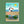 A Christmas card set wrapped in a tan paper band labeled 'ONNEKE', featuring coastal winter illustrations with a puffin, waves, a yellow sun, and green seaweed. The set is photographed on a textured olive green background.