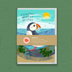 A Christmas card set wrapped in a tan paper band labeled 'ONNEKE', featuring coastal winter illustrations with a puffin, waves, a yellow sun, and green seaweed. The set is photographed on a textured olive green background.