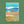 A rectangular greeting card or art print with a coastal winter illustration, shown standing against a textured green background. The artwork features turquoise and blue ocean waves, white surf, a person at the water's edge, birds in the sky, and a sandy beach area with the text 'Coastal Winter Wishes.