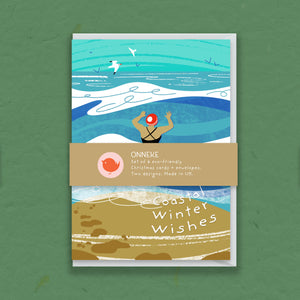 A rectangular greeting card or art print with a coastal winter illustration, shown standing against a textured green background. The artwork features turquoise and blue ocean waves, white surf, a person at the water's edge, birds in the sky, and a sandy beach area with the text 'Coastal Winter Wishes.