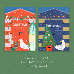 Two Christmas beach hut greeting cards shown side by side on a green textured background, with text below reading '3 of each card +6 white envelopes + belly band'. The left card has a red beach hut with a surfboard and seagull, and the right card has a navy beach hut with a decorated Christmas tree and gull.