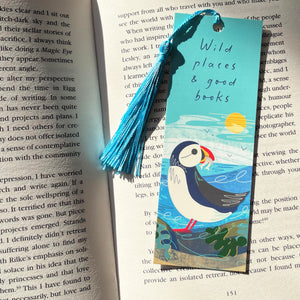 A decorative paper bookmark with a blue tassel lies on an open book page. The bookmark features a painted puffin, green seaweed, and the phrase “Wild places & good books” on a light blue background.