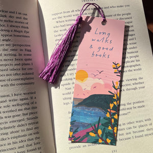 A rectangular paper bookmark with a purple tassel hangs over open book pages. The bookmark features a pastel landscape illustration with a pink sky, blue water, yellow flowers, and the handwritten text 'Long walks & good books'.