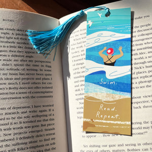A handmade paper bookmark with a blue sky and ocean illustration, featuring a swimmer and the words 'Swim. Read. Repeat.'