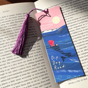 A decorative paper bookmark with a purple tassel lies on top of an open book page. The bookmark features an illustrated scene with a pink sky, full moon, dark blue water, and a red bobble hat on a swimmer, plus the text 'Dip. Dry. Read.'