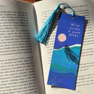 A decorative bookmark shaped like a book page with a blue tassel, featuring an illustration of ocean waves and a pink moon, placed on open book pages. The bookmark has the words 'Wild swims & good books' printed on it.