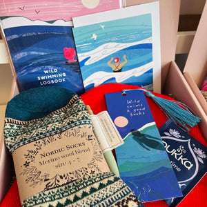 A curated gift box or flat-lay arrangement featuring a pair of patterned knit socks with a paper label reading 'Nordic Socks Merino wool blend size 4-7', illustrated swim-themed cards or booklets, a tassel bookmark, and a blue card, all arranged inside a pink box.